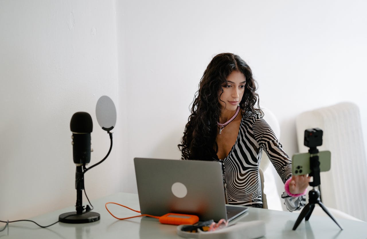 A young woman using a laptop and smartphone for livestreaming, surrounded by tech gadgets indoors.